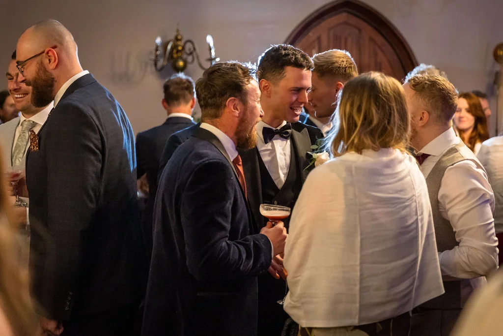 A lively wedding guests reception where family and friends mingle indoors, holding drinks and laughing during the evening celebration.
