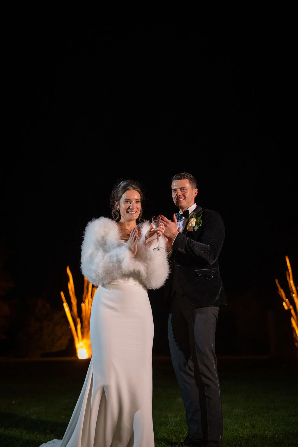 A bride and groom smile and hold a champagne glass outside during a night portrait session, with the bride wearing a white crepe gown and a fluffy white stole.