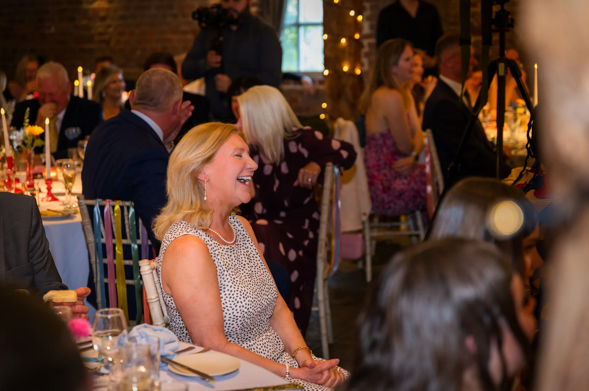 A female wedding guest in a polka dot dress laughs joyfully during the wedding reception speeches inside a rustic barn venue.