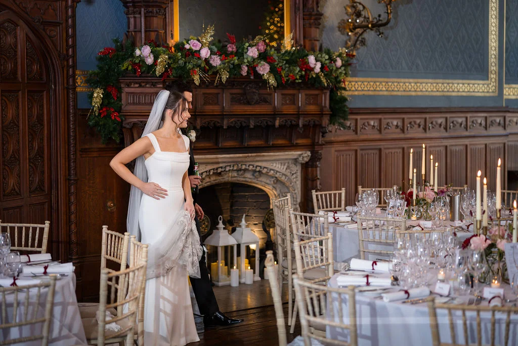A bride in a square-neckline white gown and veil stands beside a groom near an elaborately decorated fireplace inside a historic manor house for a winter wedding reception.
