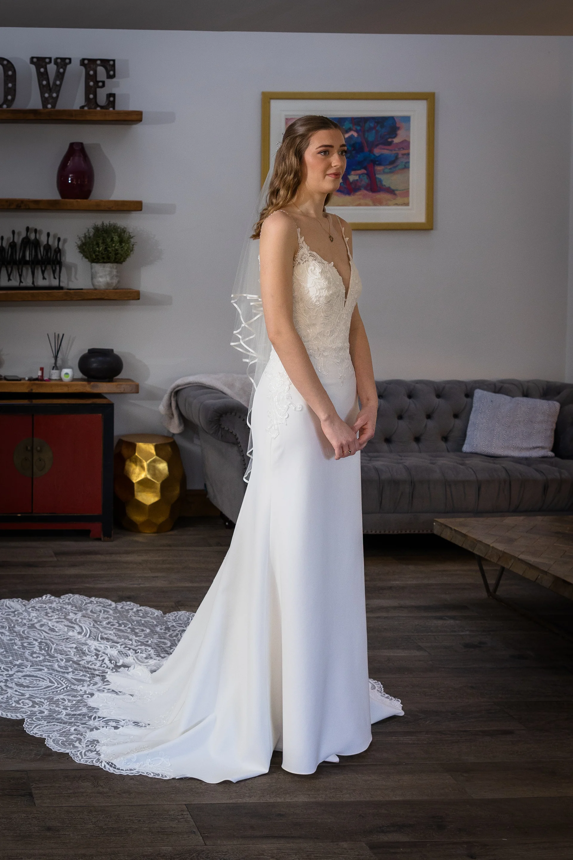 A graceful bride stands in a brightly lit room wearing a sleek crepe gown with a plunging v-neckline and a delicate lace-edged veil.