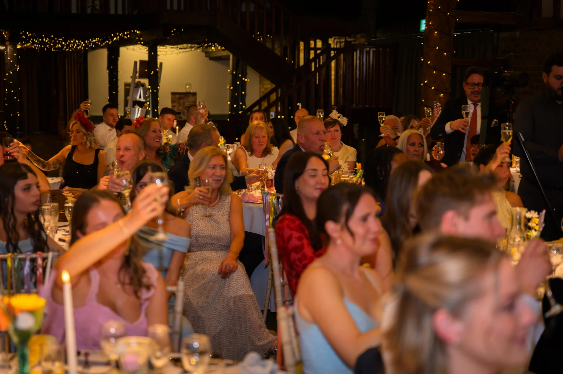 Wedding guests enjoy speeches and raise glasses of champagne during a candlelit reception inside a rustic oak-framed barn.