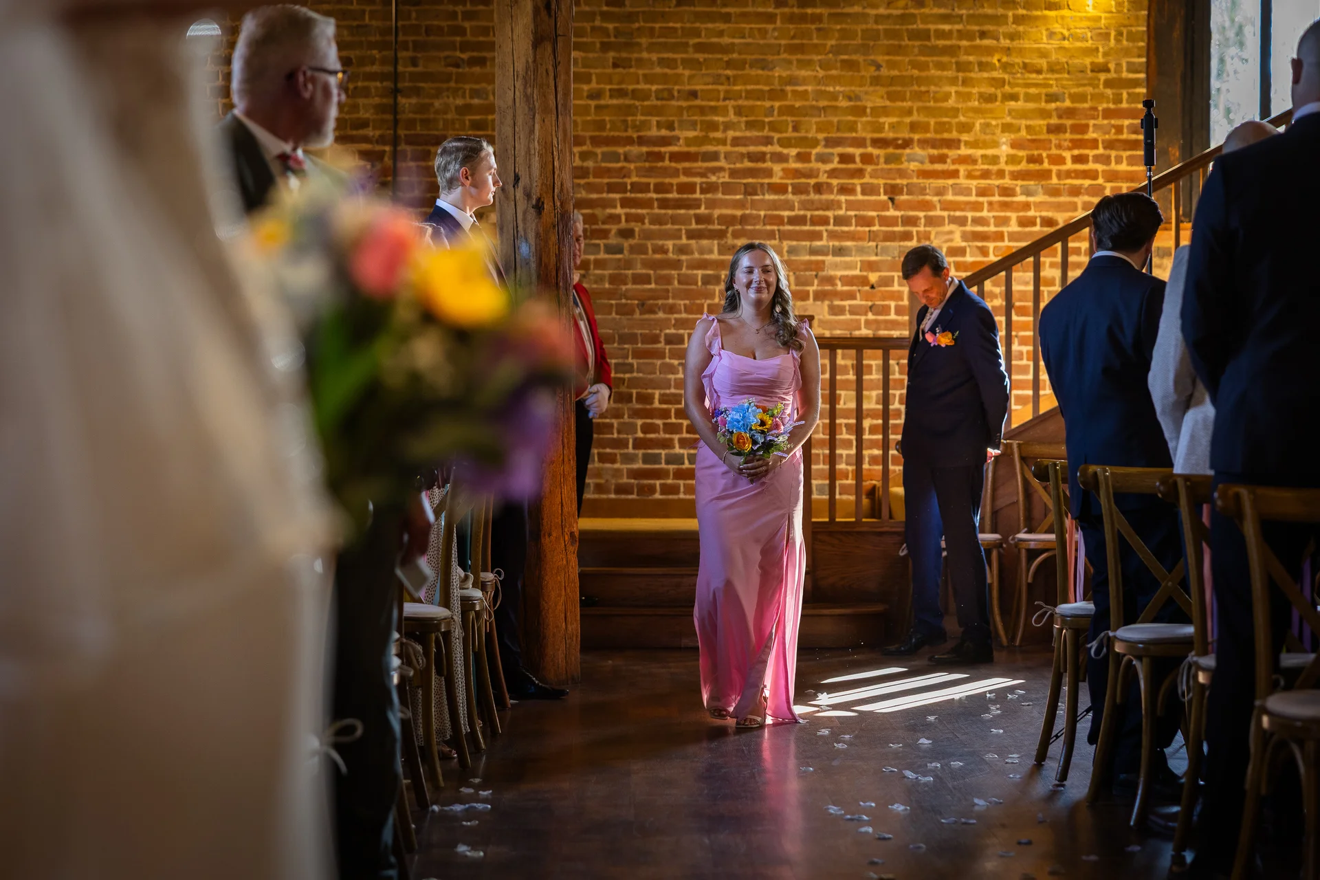 A bridesmaid in a pink dress walks down a rustic wooden aisle lined with chairs inside a brick barn, holding a colourful bouquet.