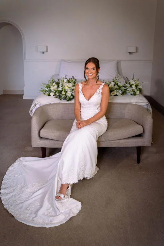 A smiling bride in a v-neck crepe gown with a sweeping lace train sits gracefully on a neutral settee with floral arrangements resting on the bed behind her.