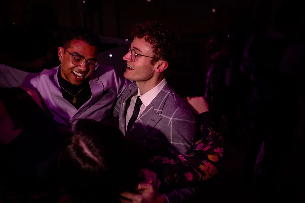 Two grooms in stylish suits laugh joyfully together on a lively, dark dance floor illuminated by vibrant purple party lights.