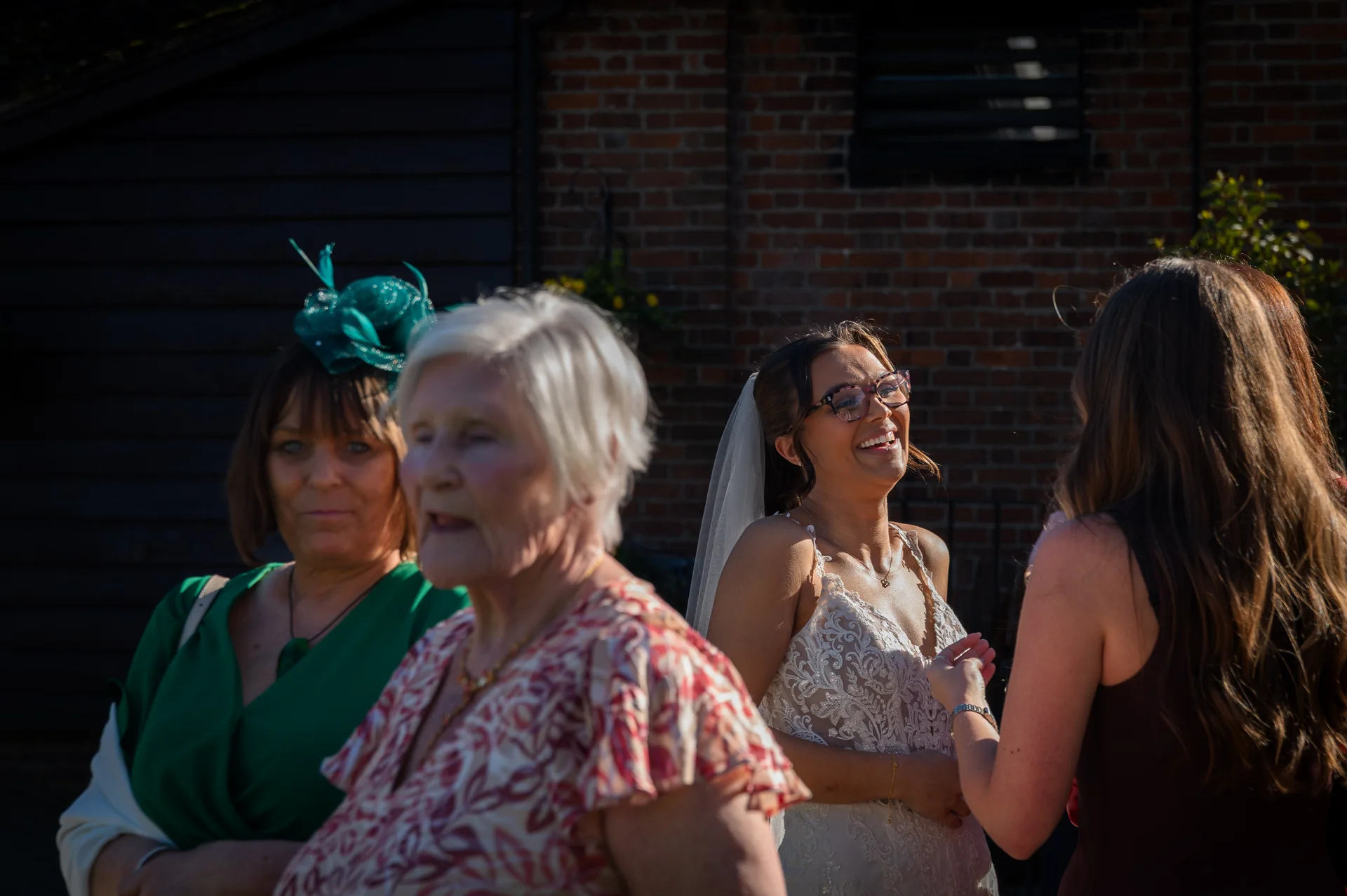 A joyful bride wearing glasses and a lace gown laughs heartily while mingling with family and guests outdoors in the bright sunshine.