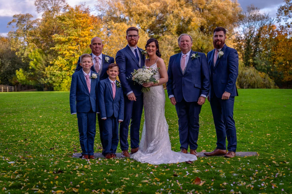 A bridal party including a bride, groom, groomsmen, and young boys poses together on a lush green lawn surrounded by vibrant autumn foliage.