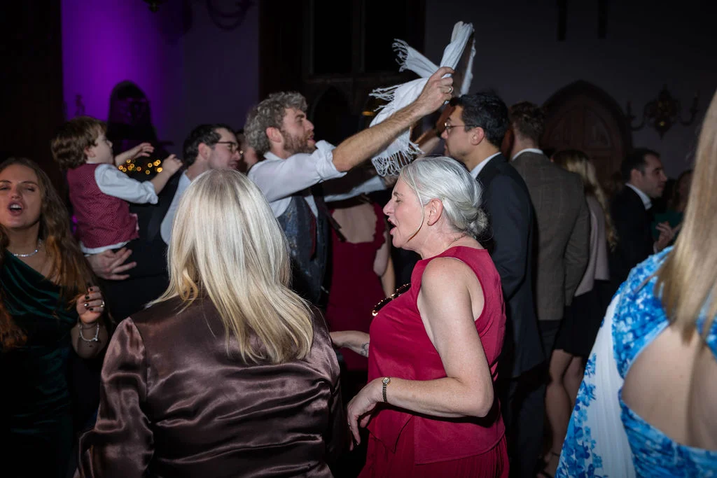 A lively group of wedding guests dance and sing enthusiastically together on a crowded evening reception dance floor.