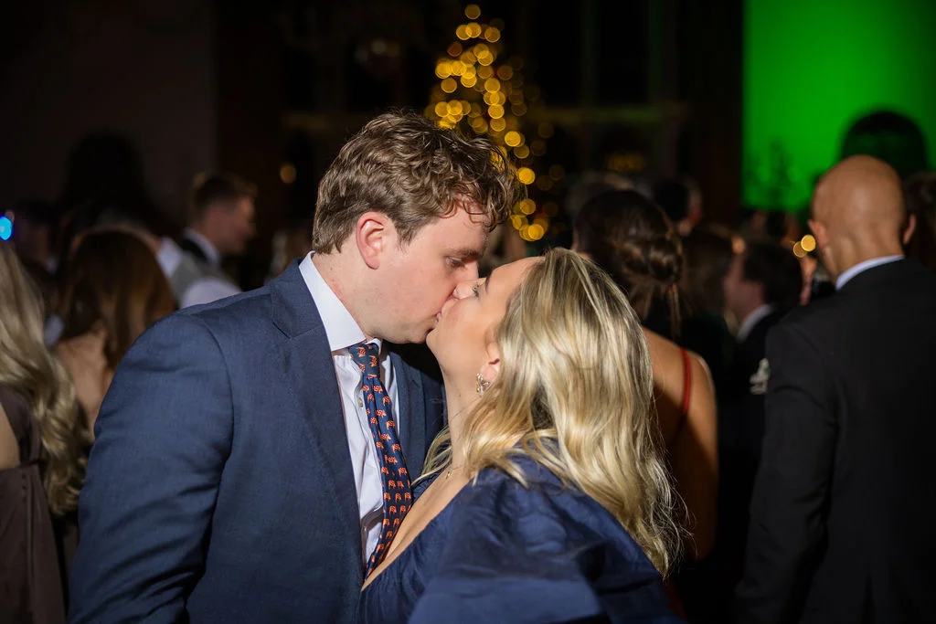 A couple shares a tender kiss on a crowded evening dance floor illuminated by warm fairy lights and vibrant green ambient lighting.