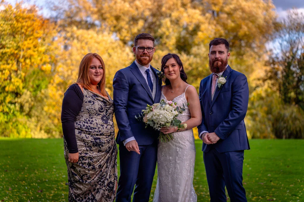 A bridal couple poses outdoors with wedding guests against a backdrop of vibrant autumn trees with golden foliage.