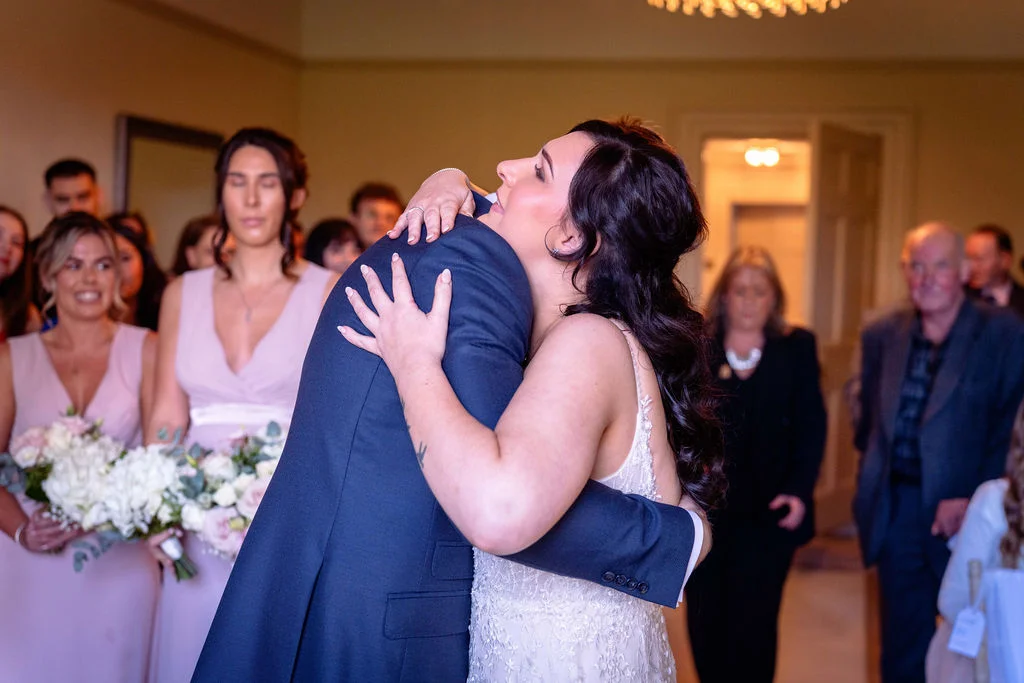 A newlywed bride in a white lace gown embraces her groom wearing a navy suit during an emotional moment, surrounded by bridesmaids in pink dresses and wedding guests.