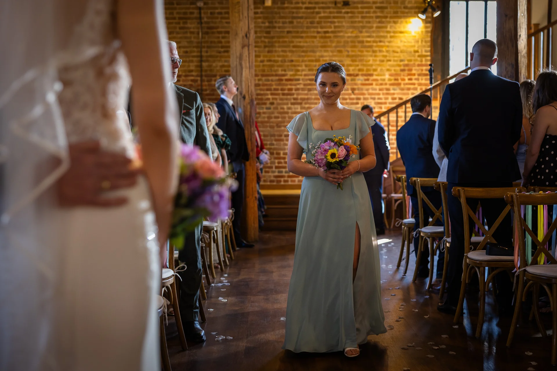 A bridesmaid in a pale blue dress with flutter sleeves and a high leg split walks down the aisle in a rustic barn venue with exposed brick walls.