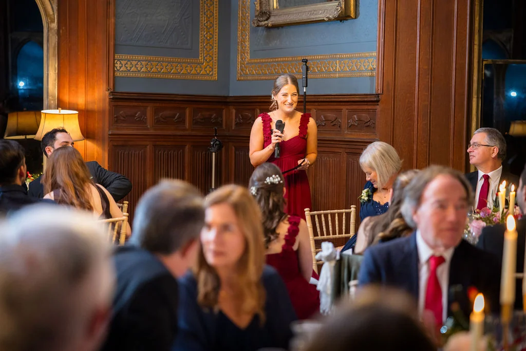 A smiling bride delivers a speech holding a microphone inside an elegant wood-panelled country house reception room filled with seated guests.