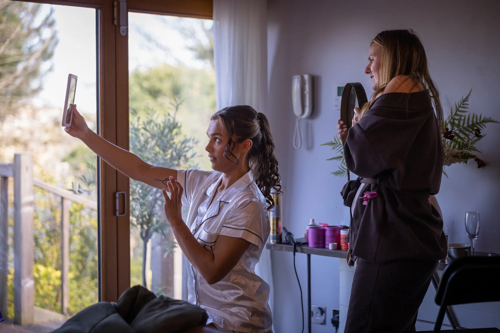 A bride in matching silk pyjamas holds up a handheld mirror by a bright window while getting ready for her wedding morning.
