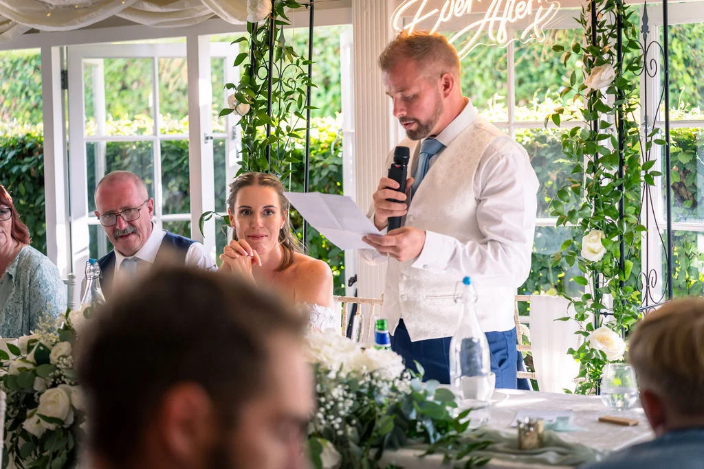 The groom delivers his wedding speech at the top table inside a bright conservatory adorned with white roses and a neon sign.
