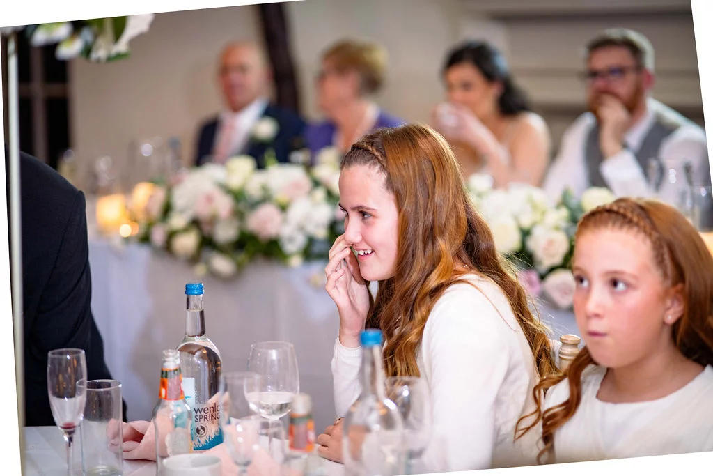 A young bridesmaid smiles and covers her mouth while seated at the top table during wedding speeches, with other guests visible in the background.