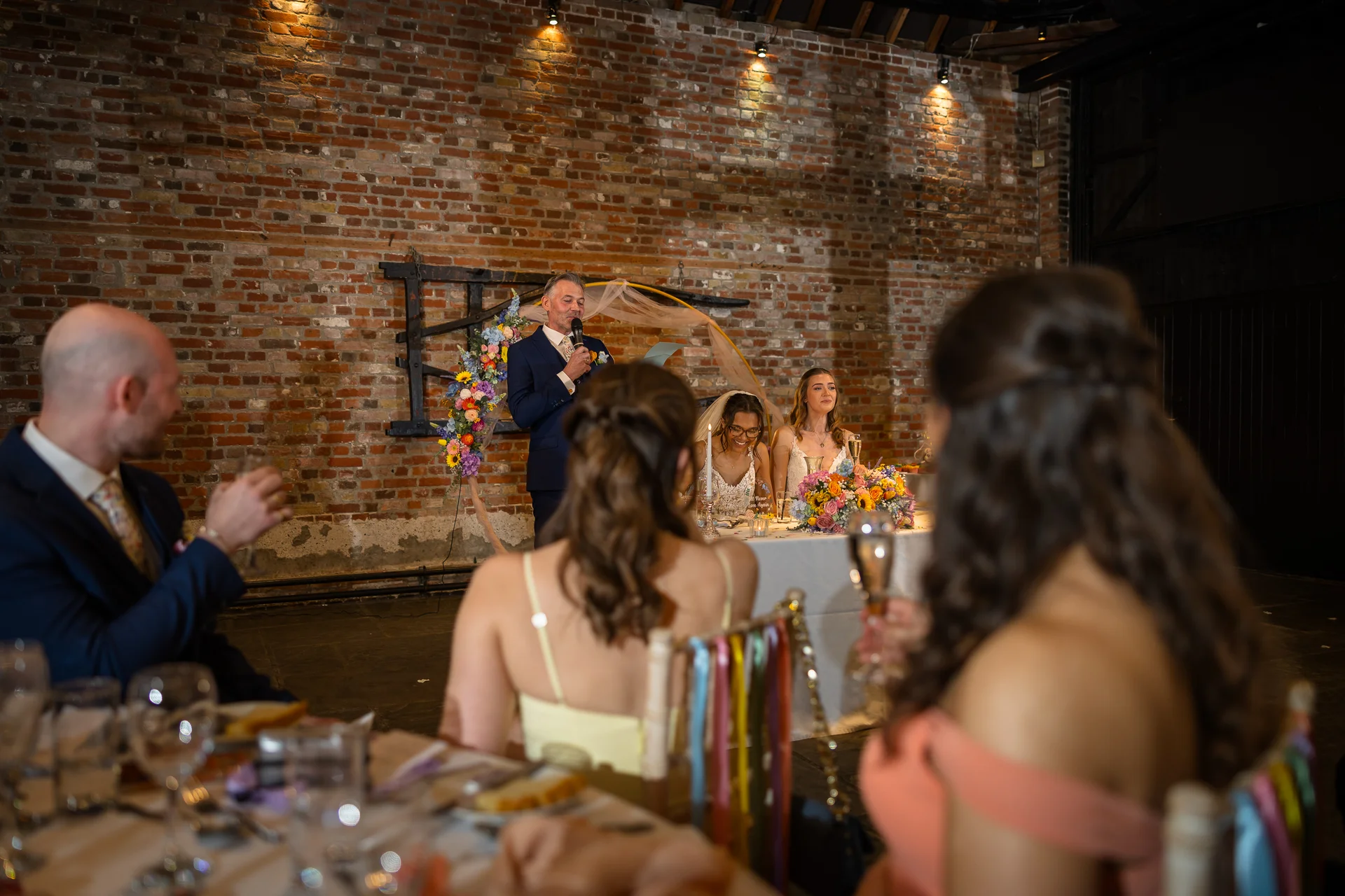 A guest delivers a speech with a microphone at the top table in an industrial barn wedding venue featuring exposed brick walls, while the smiling couple and guests listen.