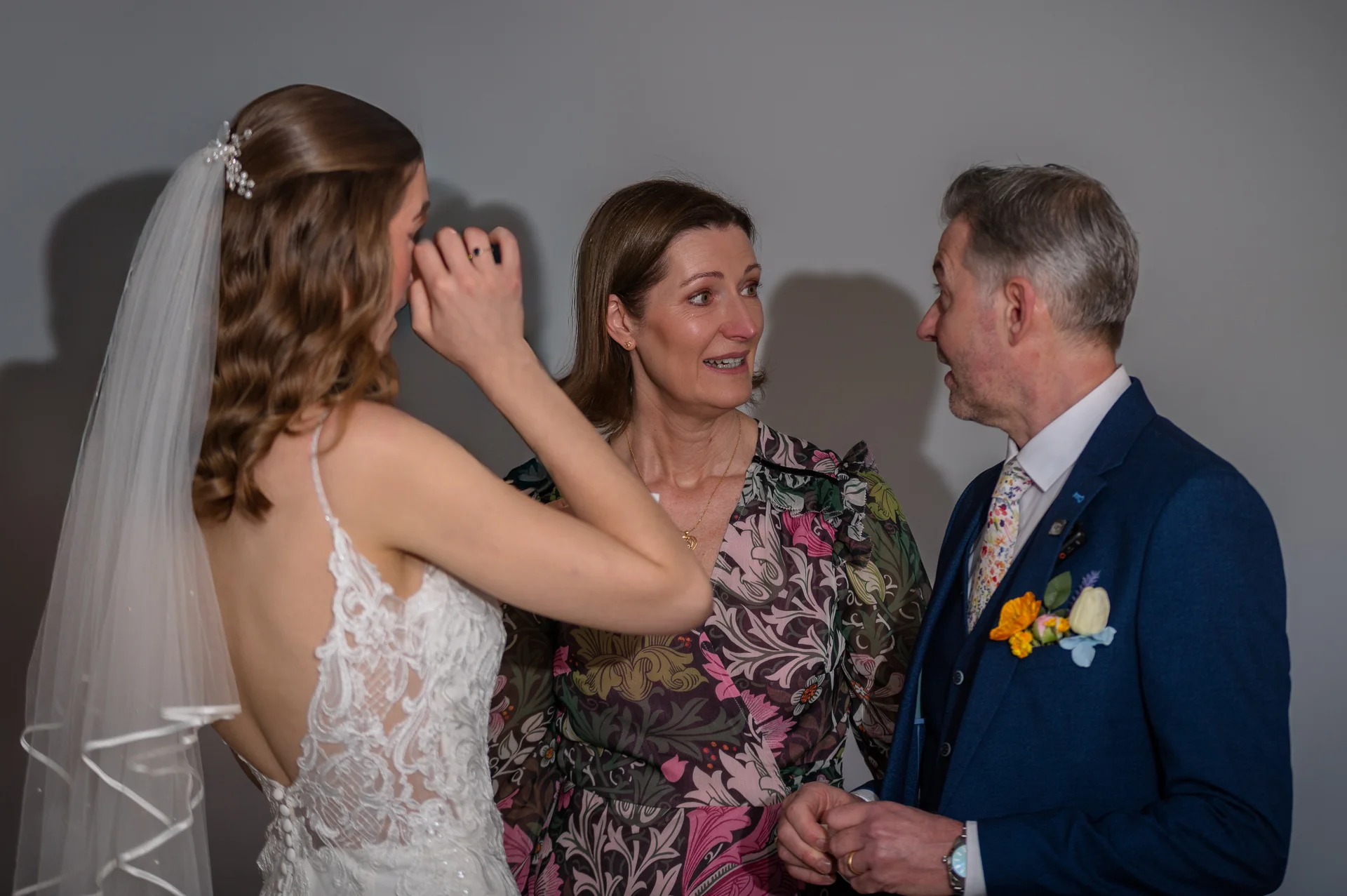 An emotional bride in a lace wedding dress with a veil wipes a tear as she shares a touching moment with her mother and father.