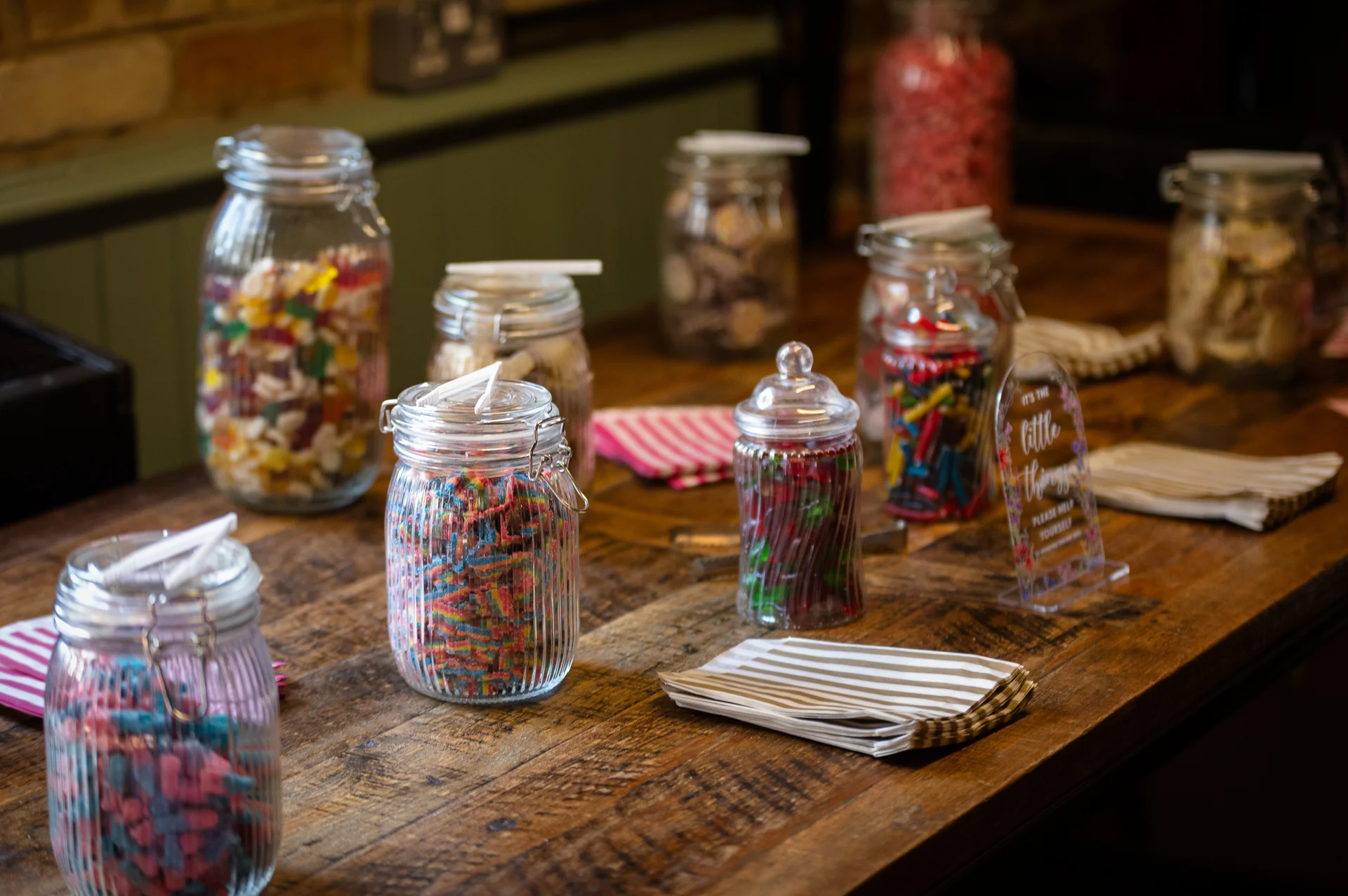 An array of glass jars filled with colorful pick and mix sweets sits on a rustic wooden table alongside striped paper bags.
