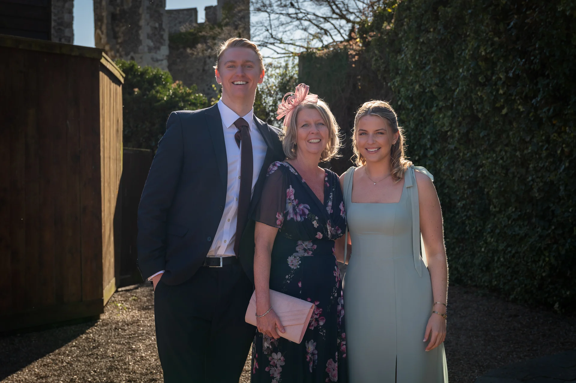 A smiling young man in a dark suit stands next to an older woman wearing a floral dress and fascinator and a younger woman in a sage green dress outdoors in the sunshine.