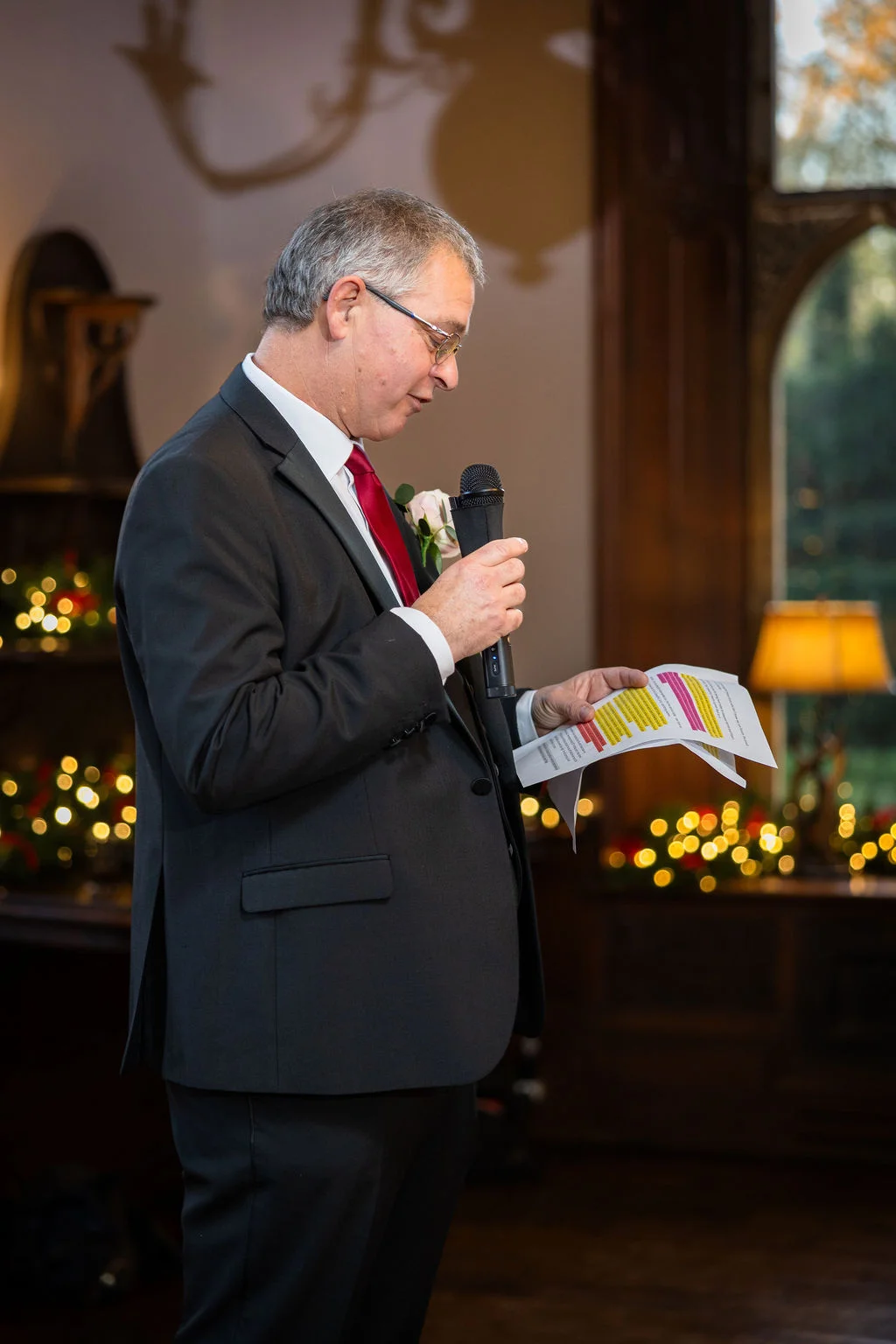 An older gentleman wearing a grey suit and a red tie holds a microphone and reads his speech from printed papers during the wedding reception.