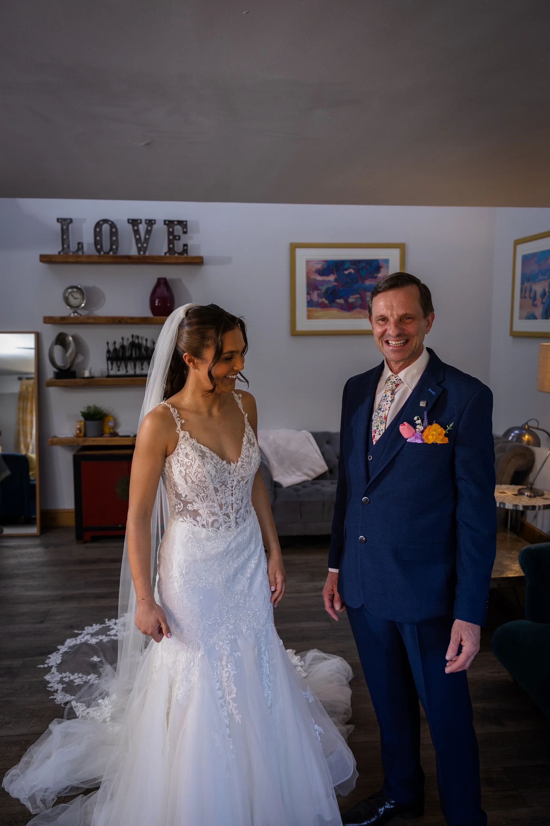 A smiling bride in a lace fit-and-flare wedding dress with a sheer corset bodice and veil shares a touching first look moment with her father in a blue suit.
