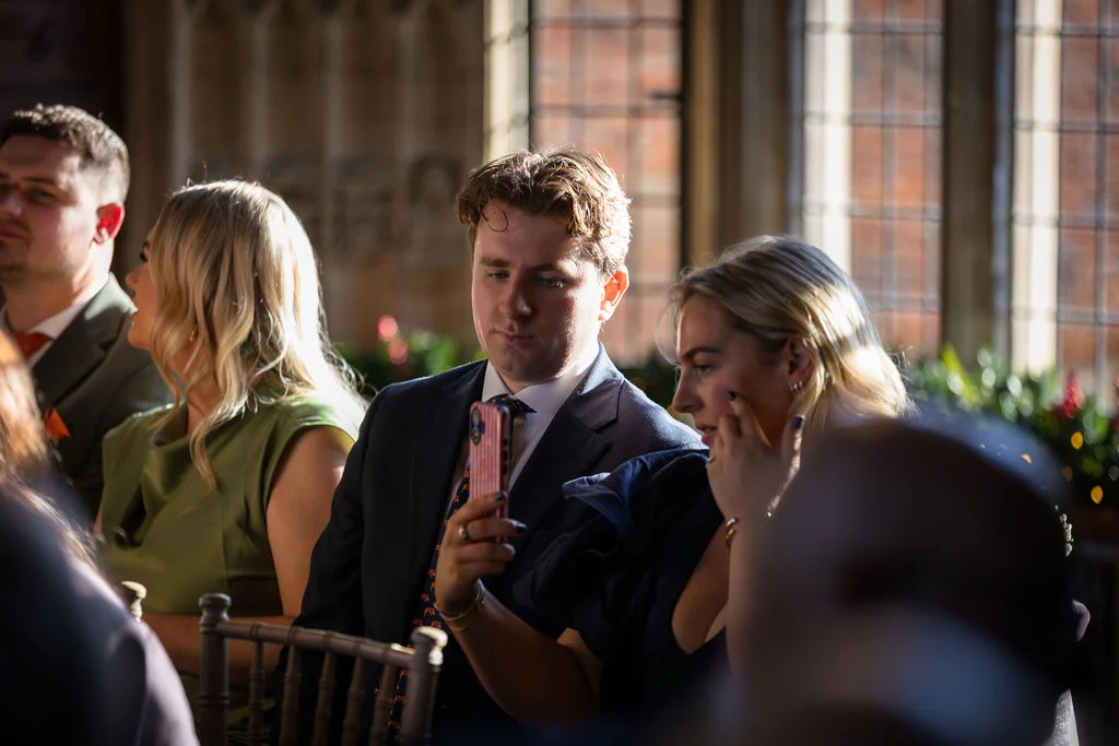 Wedding guests sit near large windows inside a historic venue, with a guest in a suit taking a photo on his phone while others look on.