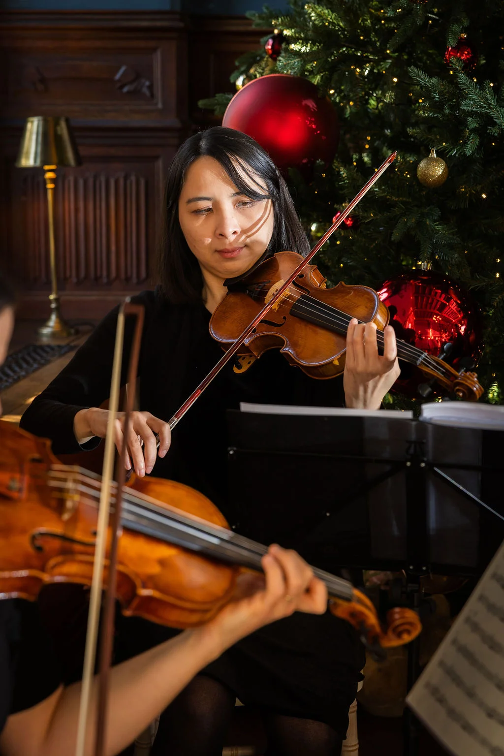 A focused female violinist plays her instrument passionately in front of a warmly lit Christmas tree adorned with red and gold baubles during a festive wedding celebration.
