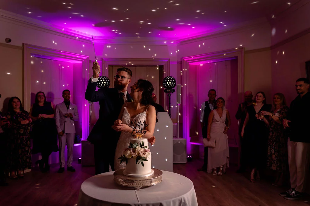 The bride and groom cut their two-tier wedding cake together while surrounded by cheering guests under a ceiling lit with vibrant purple disco lights.