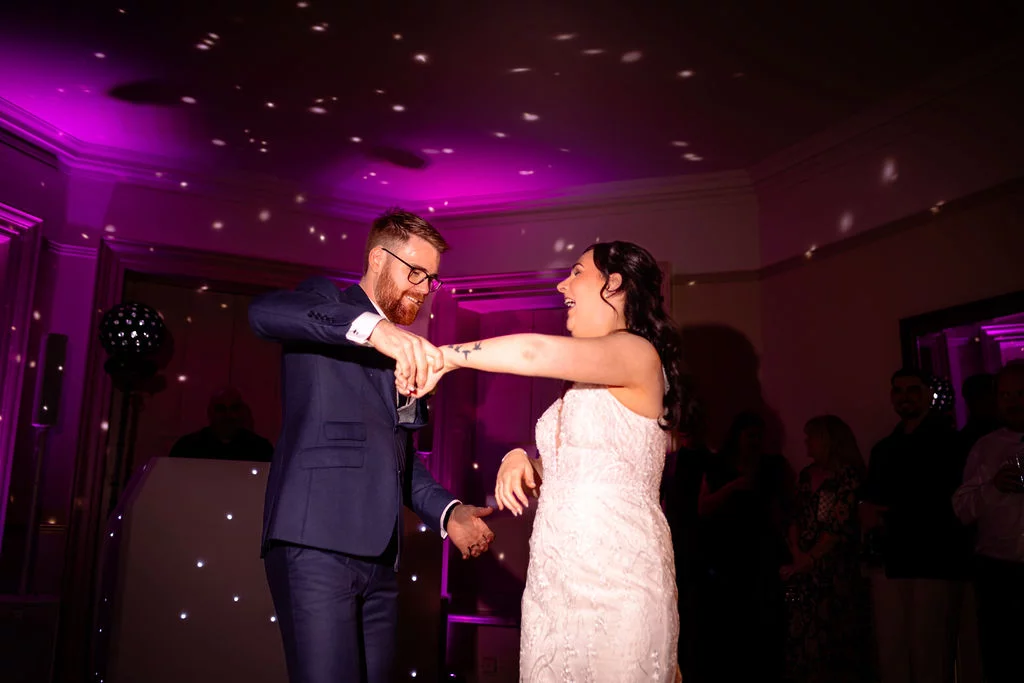 A newlywed couple share a joyful first dance under vibrant pink lighting and sparkling reflections on the ceiling during their evening reception.