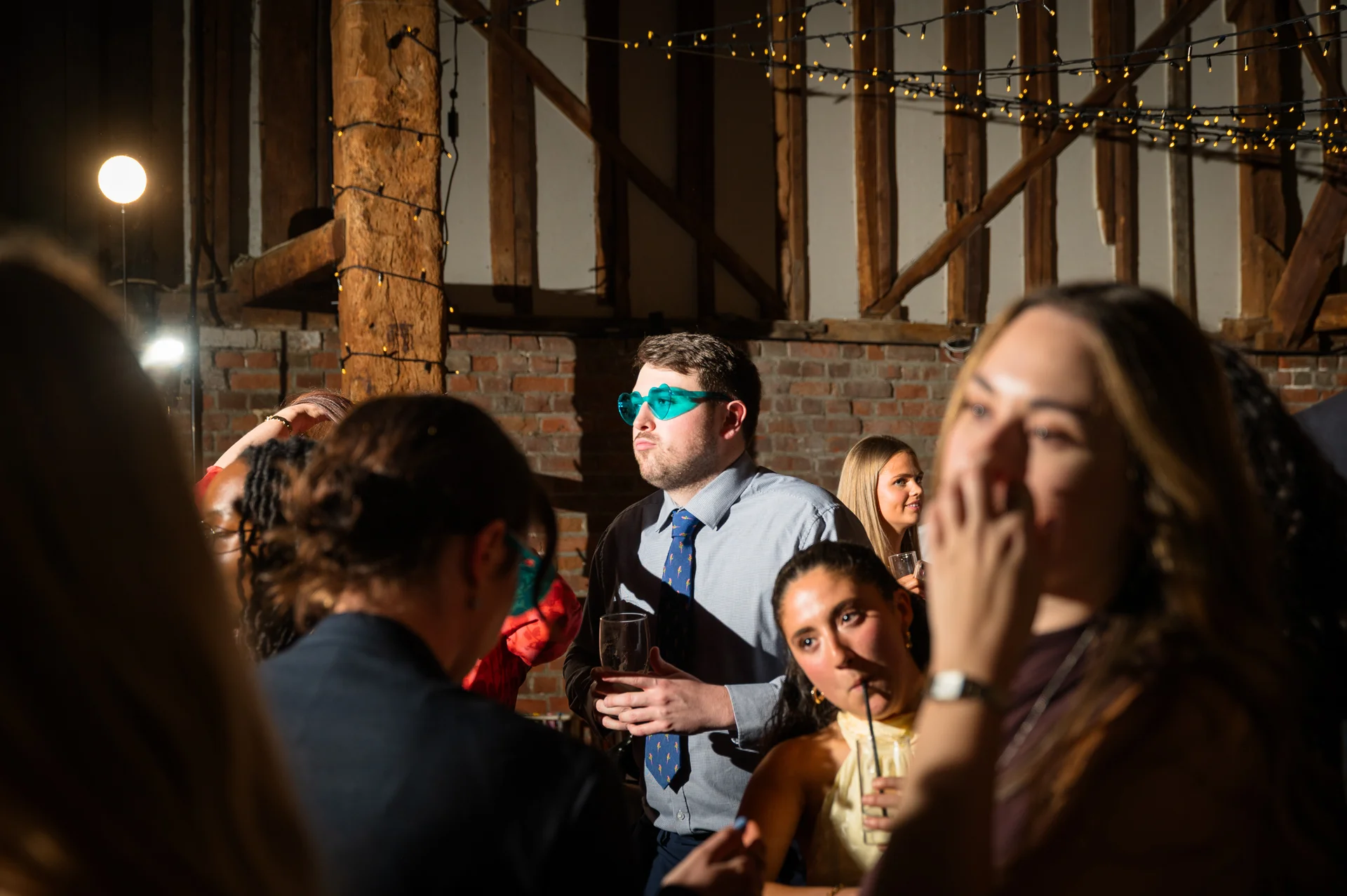 A lively evening wedding reception inside a rustic barn featuring exposed wooden beams, brick walls, and festoon lighting where guests mingle and enjoy drinks.