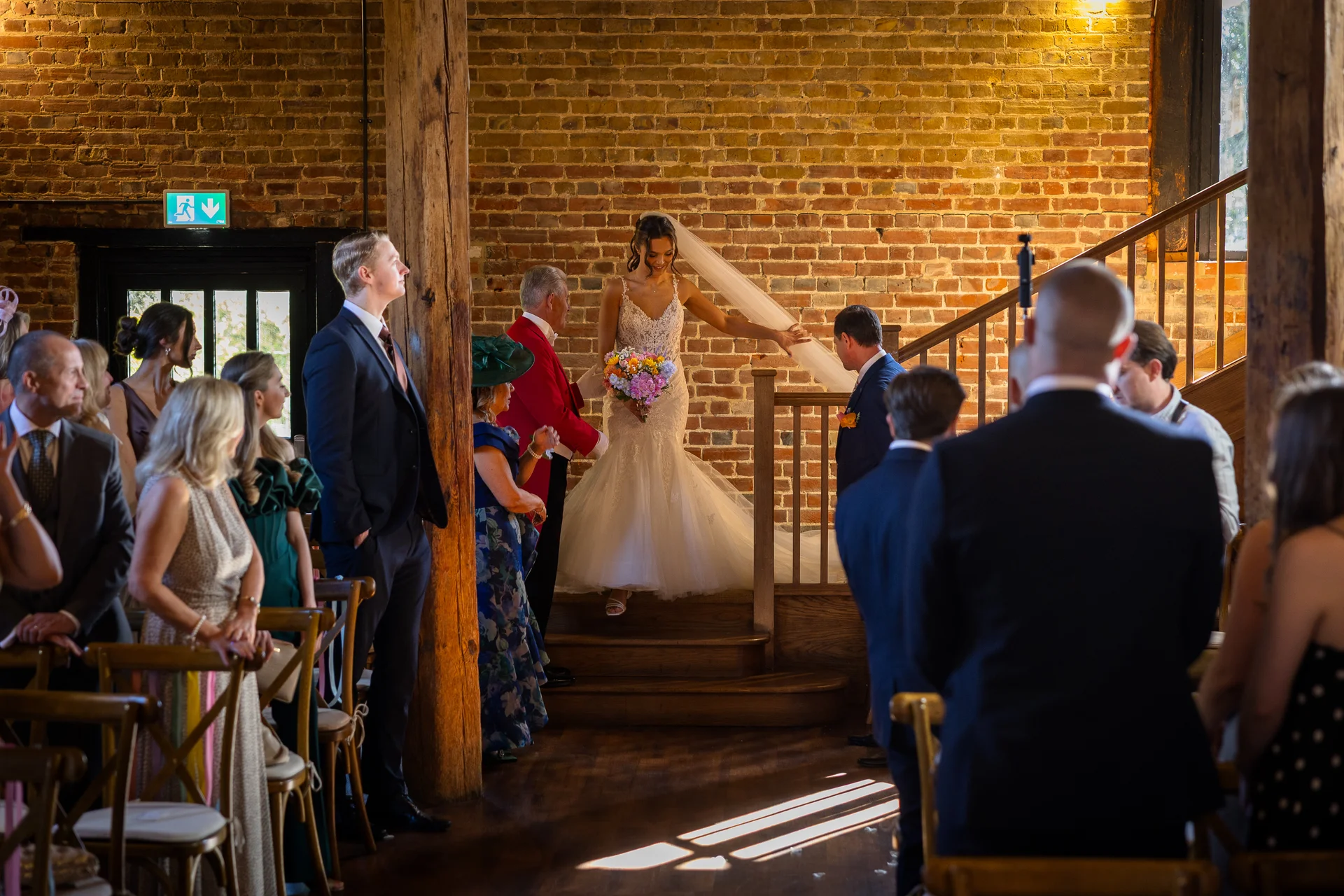 A smiling bride holding a colourful bouquet descends a wooden staircase inside a rustic stone and brick barn, surrounded by wedding guests.