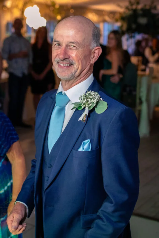 A smiling mature groom wearing a navy three-piece suit, light blue tie, and a matching buttonhole featuring baby's breath at a wedding reception.