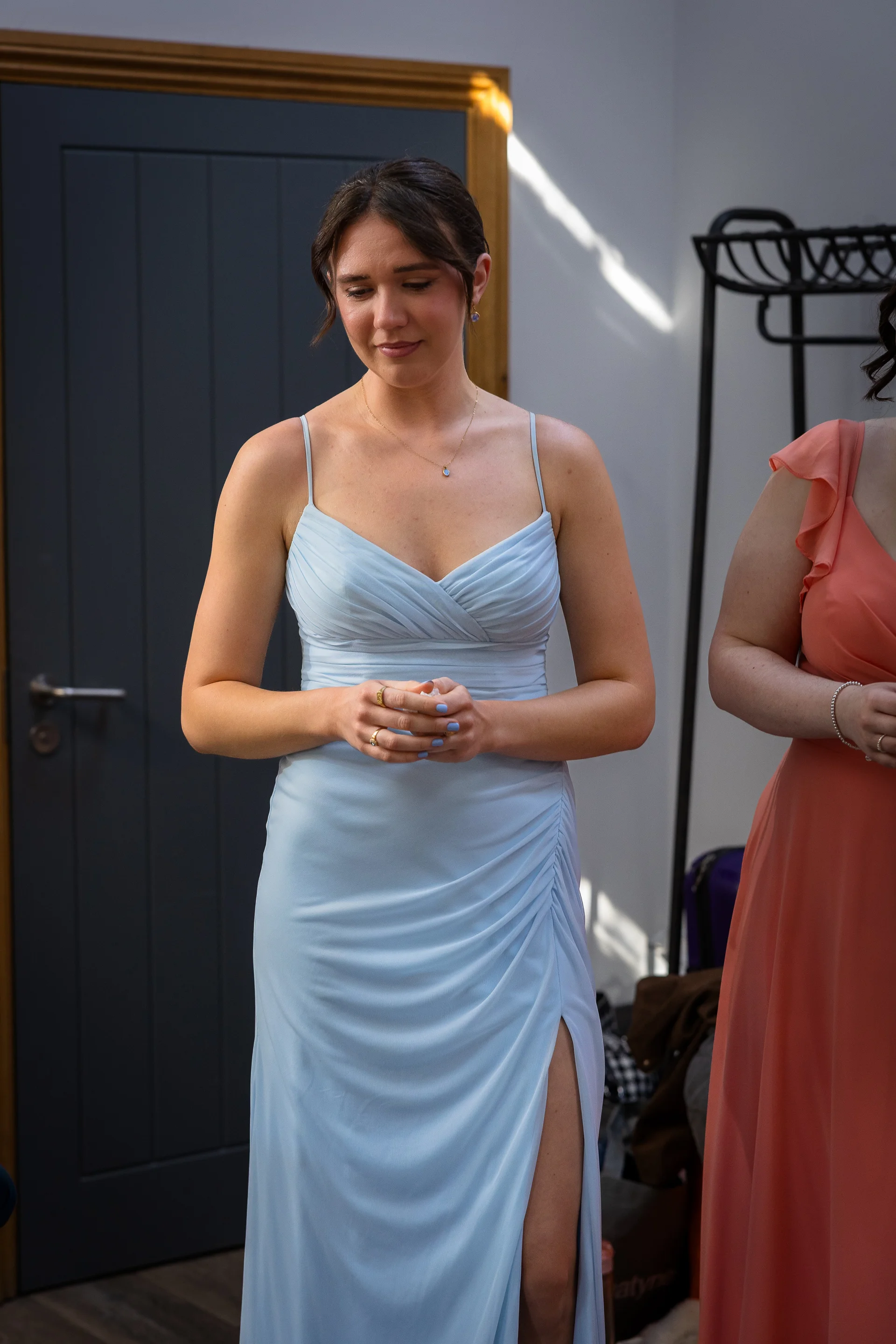 A bridesmaid wearing a light blue ruched gown with a thigh split looks down thoughtfully while holding a small tissue during morning preparations.