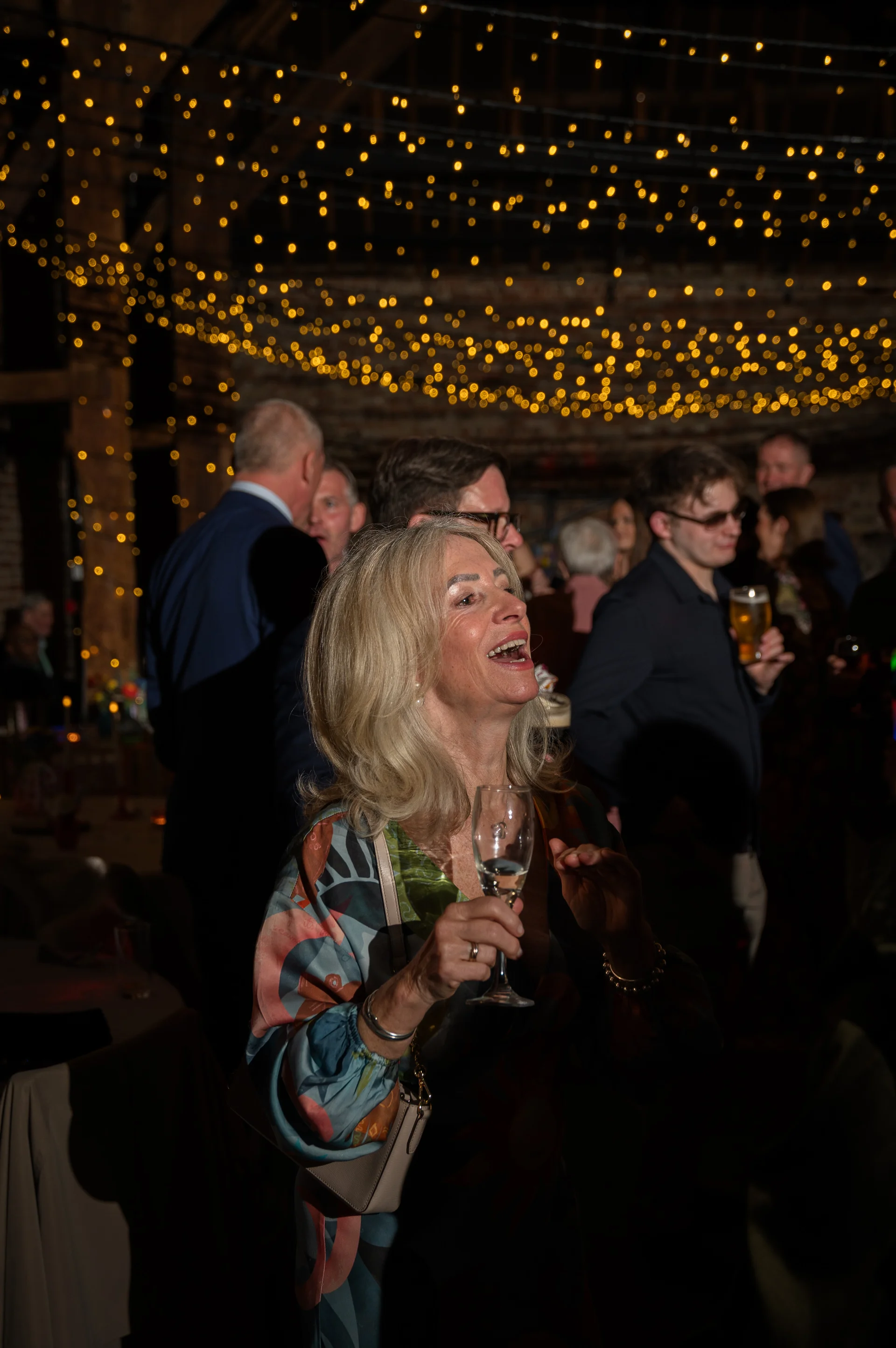 A smiling wedding guest holds a glass of champagne, laughing amidst a lively crowd beneath a glowing canopy of fairy lights inside a rustic barn venue.
