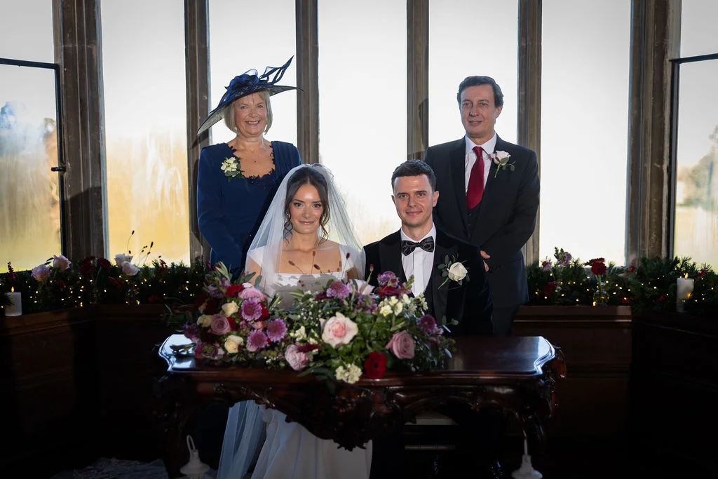 A newlywed couple poses behind a floral-decorated wooden table with their parents standing proudly beside them in a grand room with large windows.
