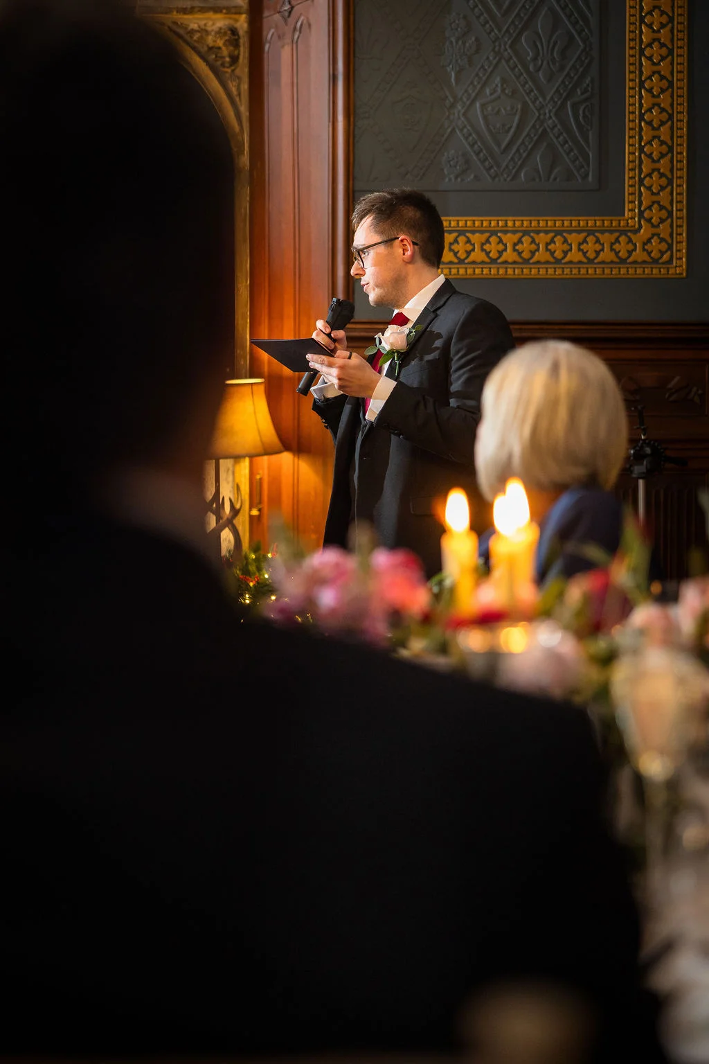 A groom wearing glasses delivers his wedding speech at a candlelit reception table while holding a microphone and notes.