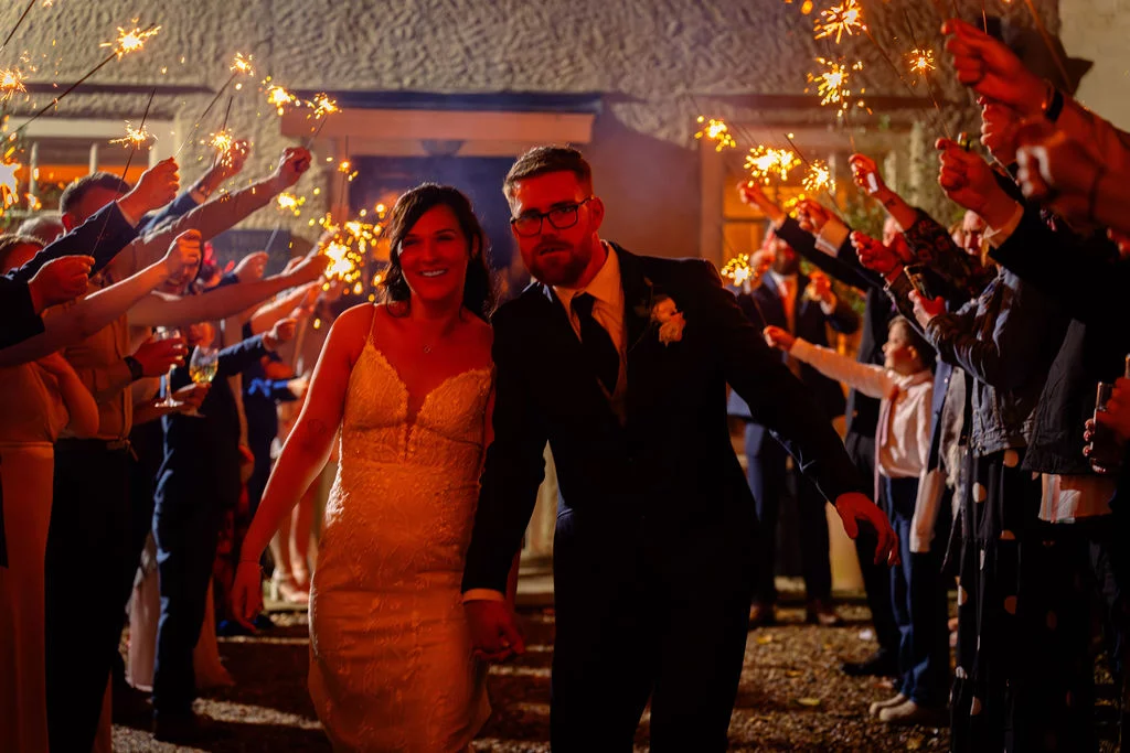 A smiling bride and groom walk hand in hand through a glowing tunnel of sparklers held by wedding guests at night.