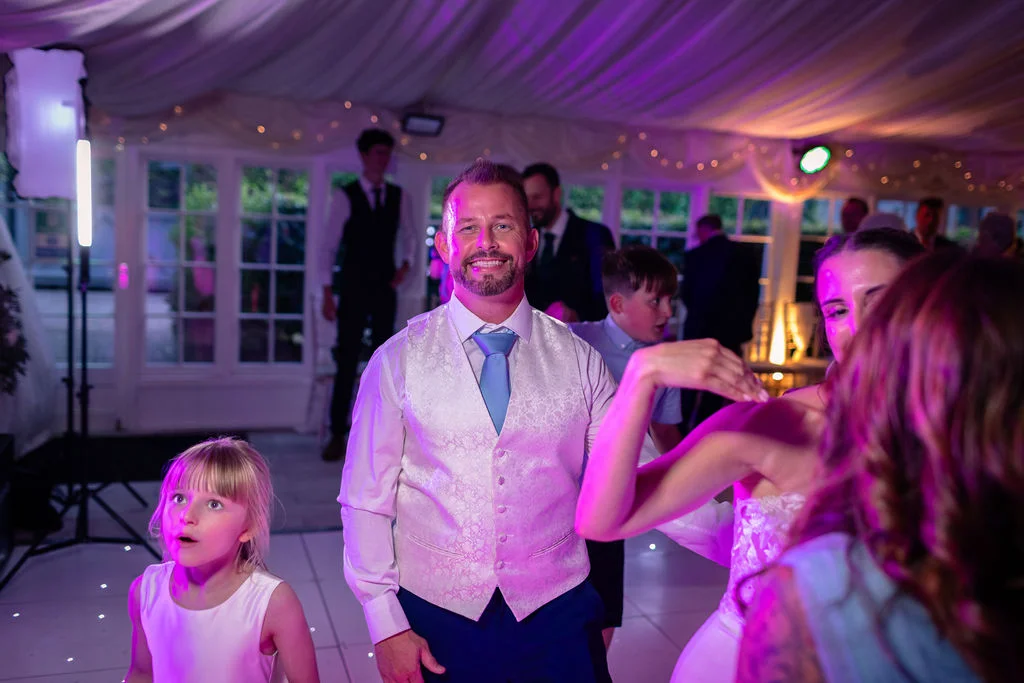 A groom smiling on the illuminated dance floor of a marquee evening reception, surrounded by wedding guests and children enjoying the celebrations under purple lighting.