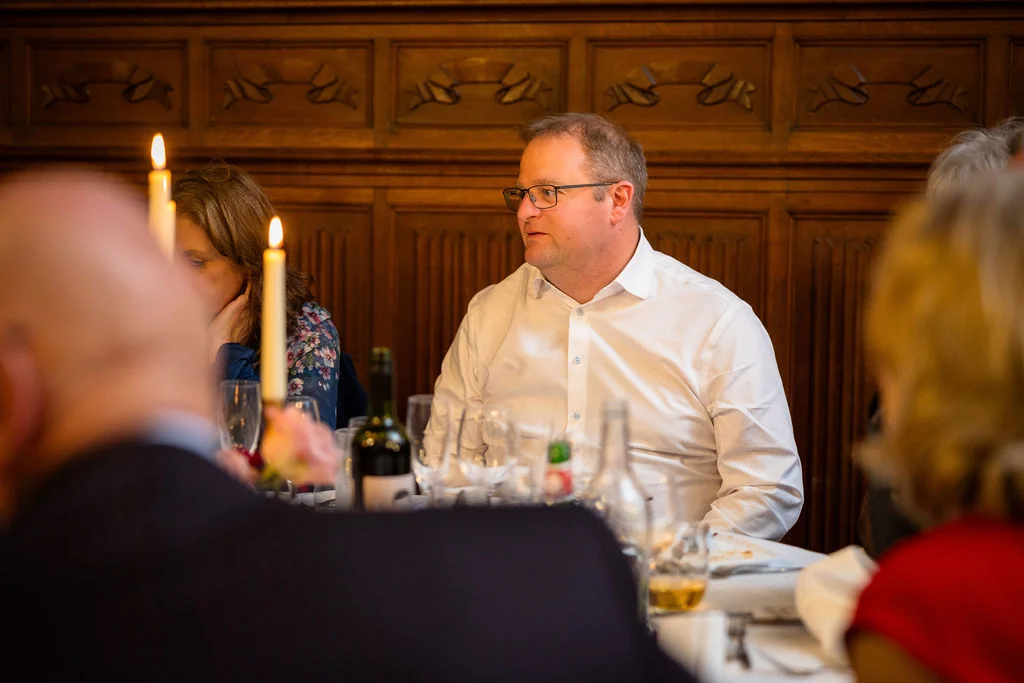 A wedding guest in a white shirt seated at a candlelit reception table during the wedding breakfast in a wood-panelled room.