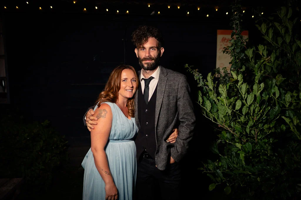 A newlywed couple poses together at night outdoors, with the groom wearing a tweed suit and black tie and the bride wearing a light blue dress with tattoos visible on her arm.