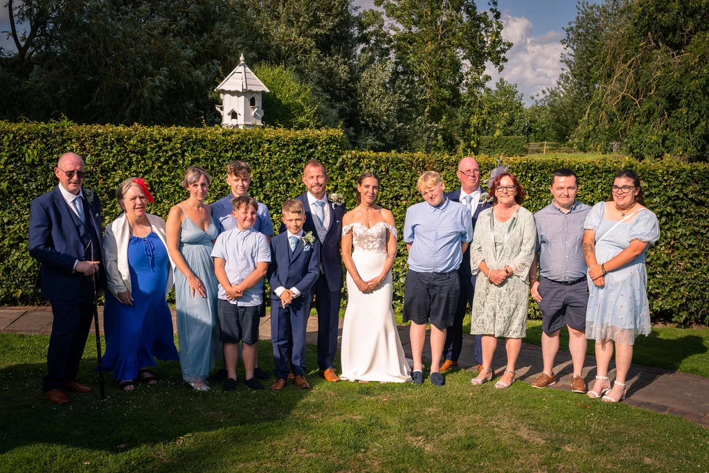 A large wedding party and family group pose together outdoors on a sunlit lawn in front of a tall green hedge.