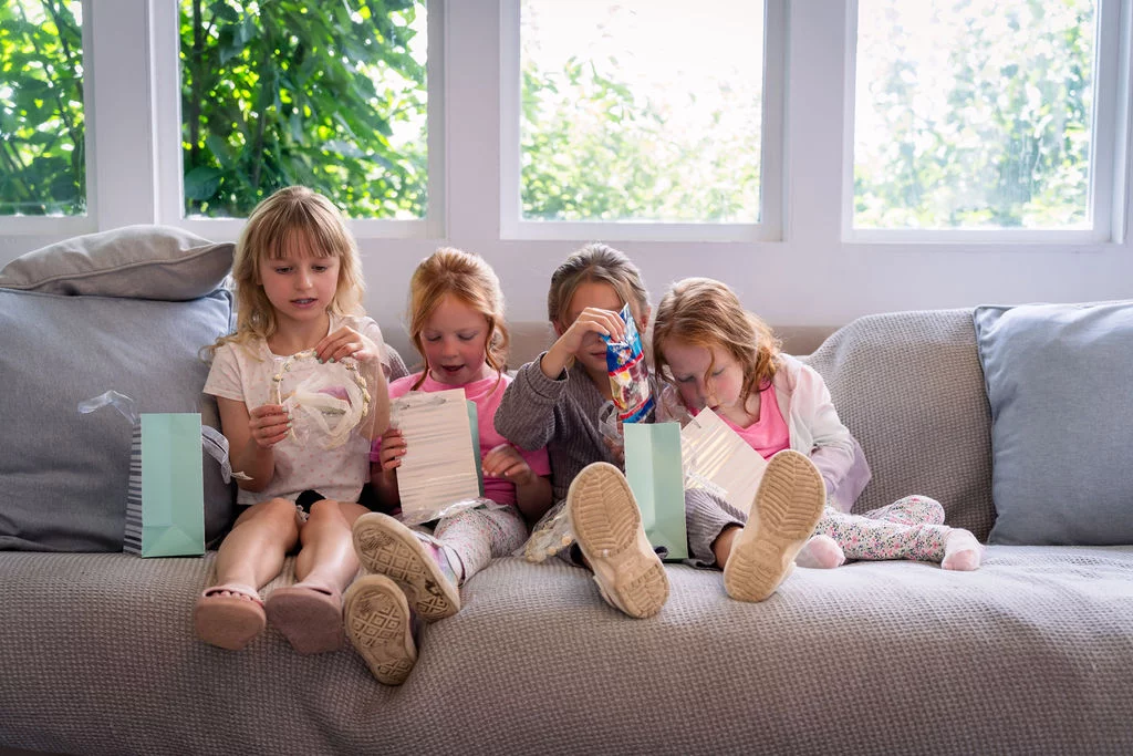 Four young flower girls happily open gift bags and inspect accessories while sitting together on a grey sofa indoors.
