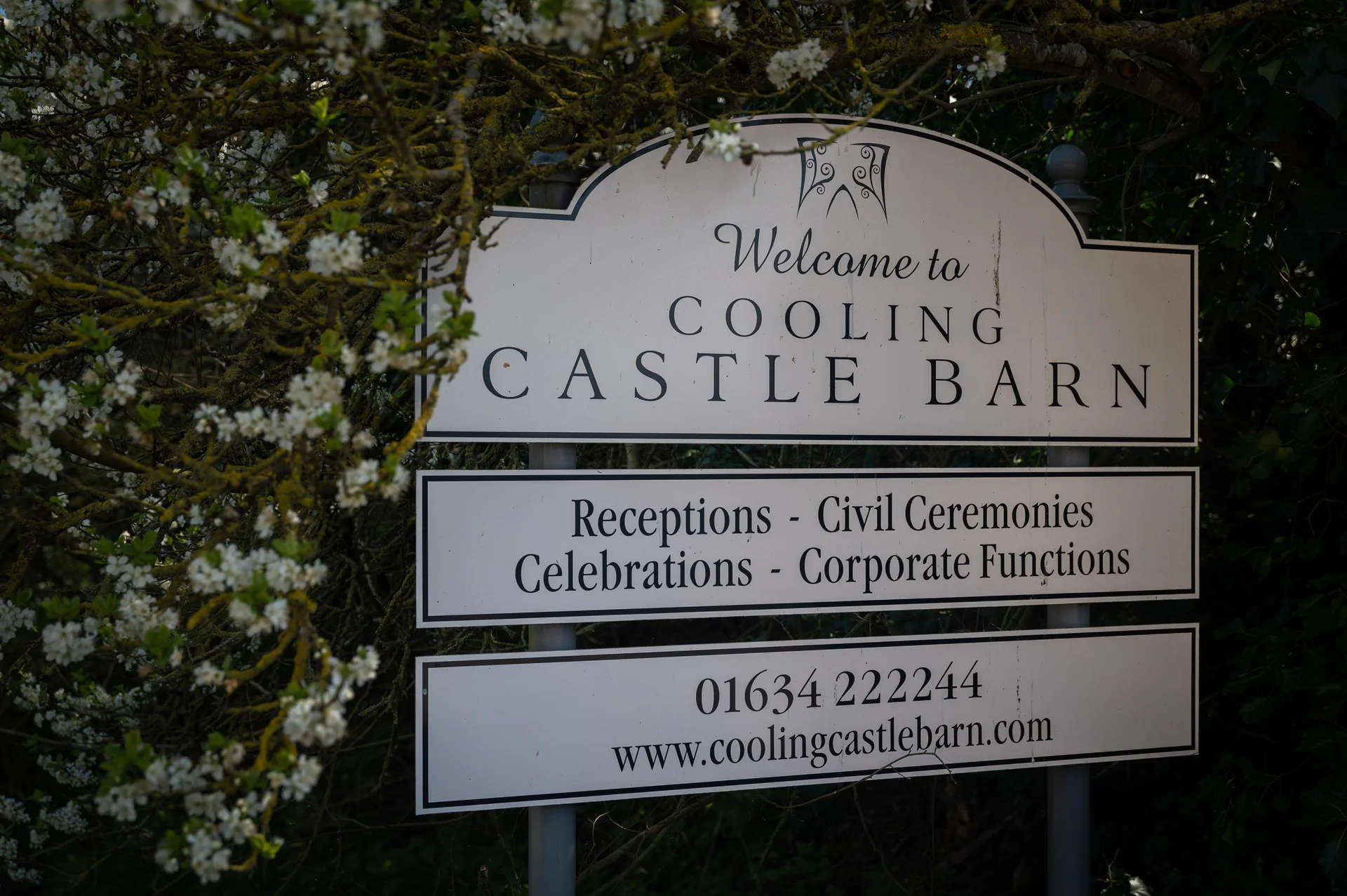 A white welcome sign for Cooling Castle Barn is partially framed by blossoming spring tree branches with white flowers.