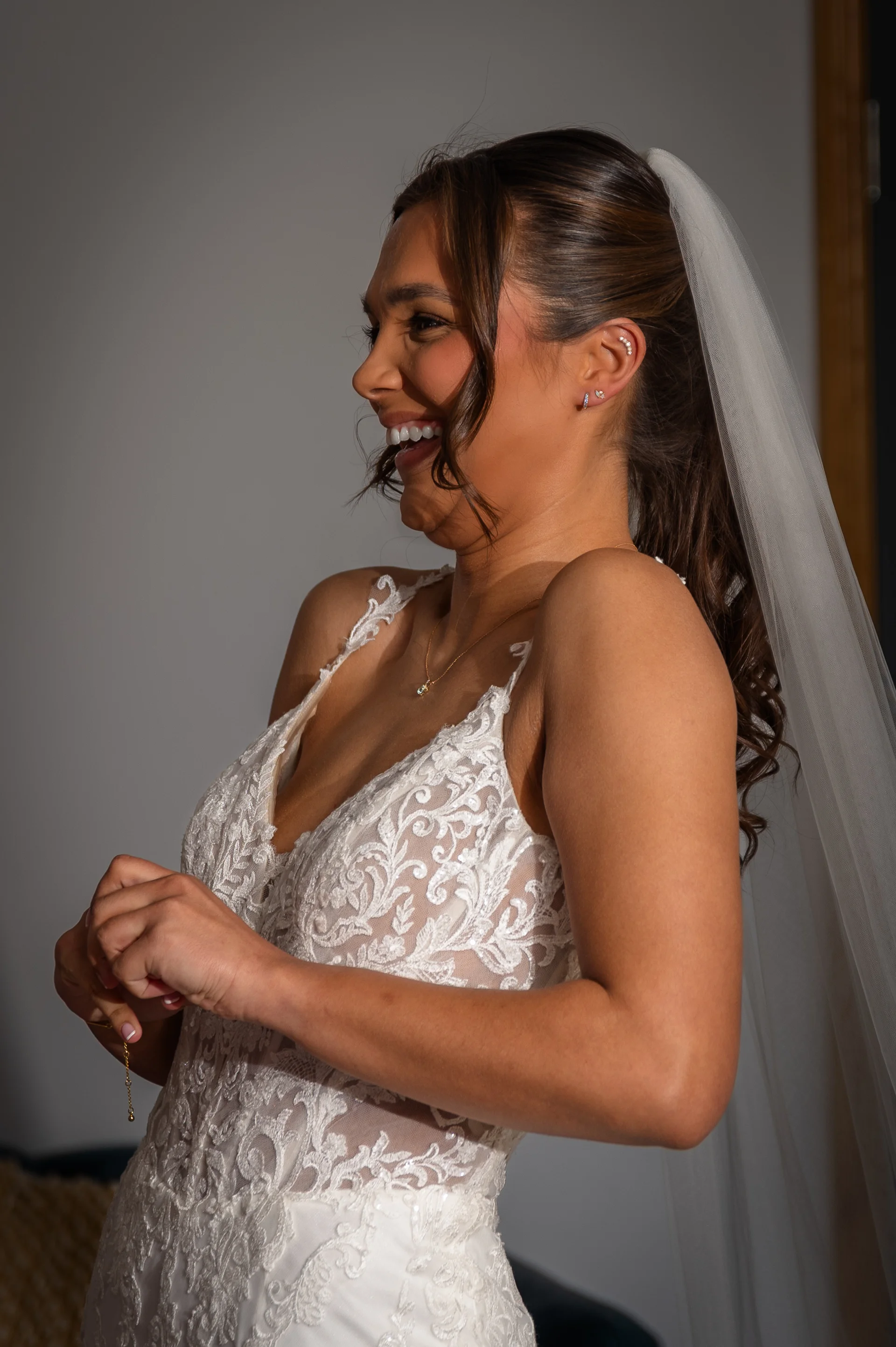 A laughing bride wearing an embroidered lace wedding dress with a deep V-neckline and a high ponytail with a veil during morning preparations.