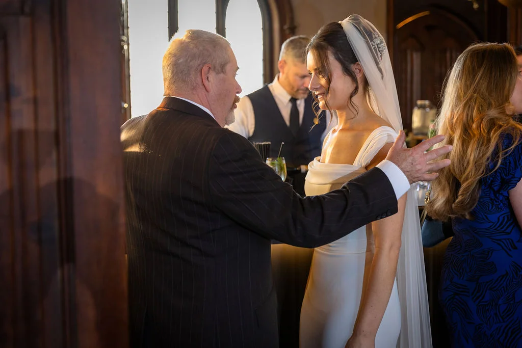 An emotional father of the bride places a tender hand on his daughter's shoulder as she smiles in her elegant satin wedding gown and veil.