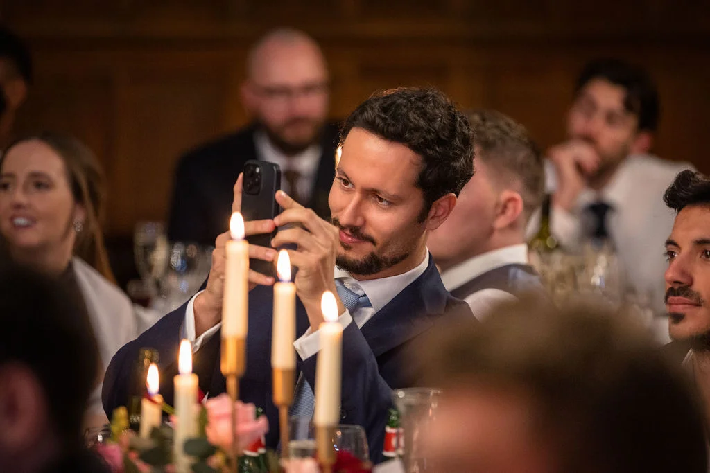 A guest at a candlelit wedding reception takes photos with his smartphone while seated at a banquet table.
