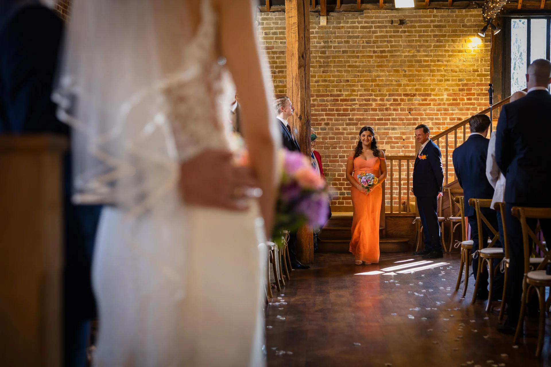 An over-the-shoulder shot captures a bridesmaid in an orange dress walking down the aisle inside a rustic brick barn wedding venue.