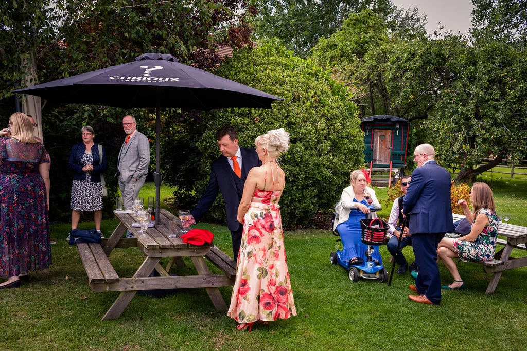 Wedding guests mingle outdoors in a lush garden setting next to wooden picnic tables and a large parasol.