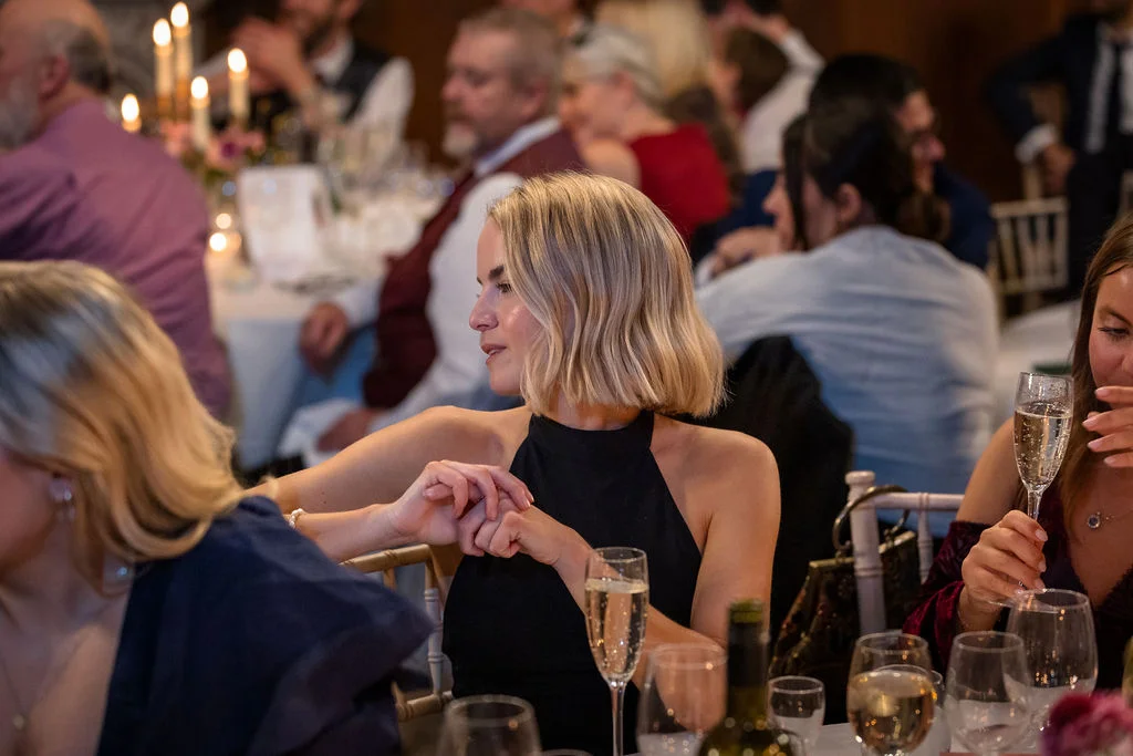 Guests are seated at a wedding breakfast, with a woman in a black halter-neck dress smiling during dinner.